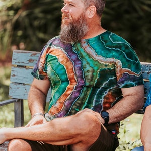 K&ouml;nnte beinhalten: Ein Mann mit langem, grauem Bart sitzt auf einer Holzbank und tr&auml;gt ein gr&uuml;n-orange-violettes Batik-Shirt und braune Shorts. Er tr&auml;gt eine Uhr am linken Handgelenk.