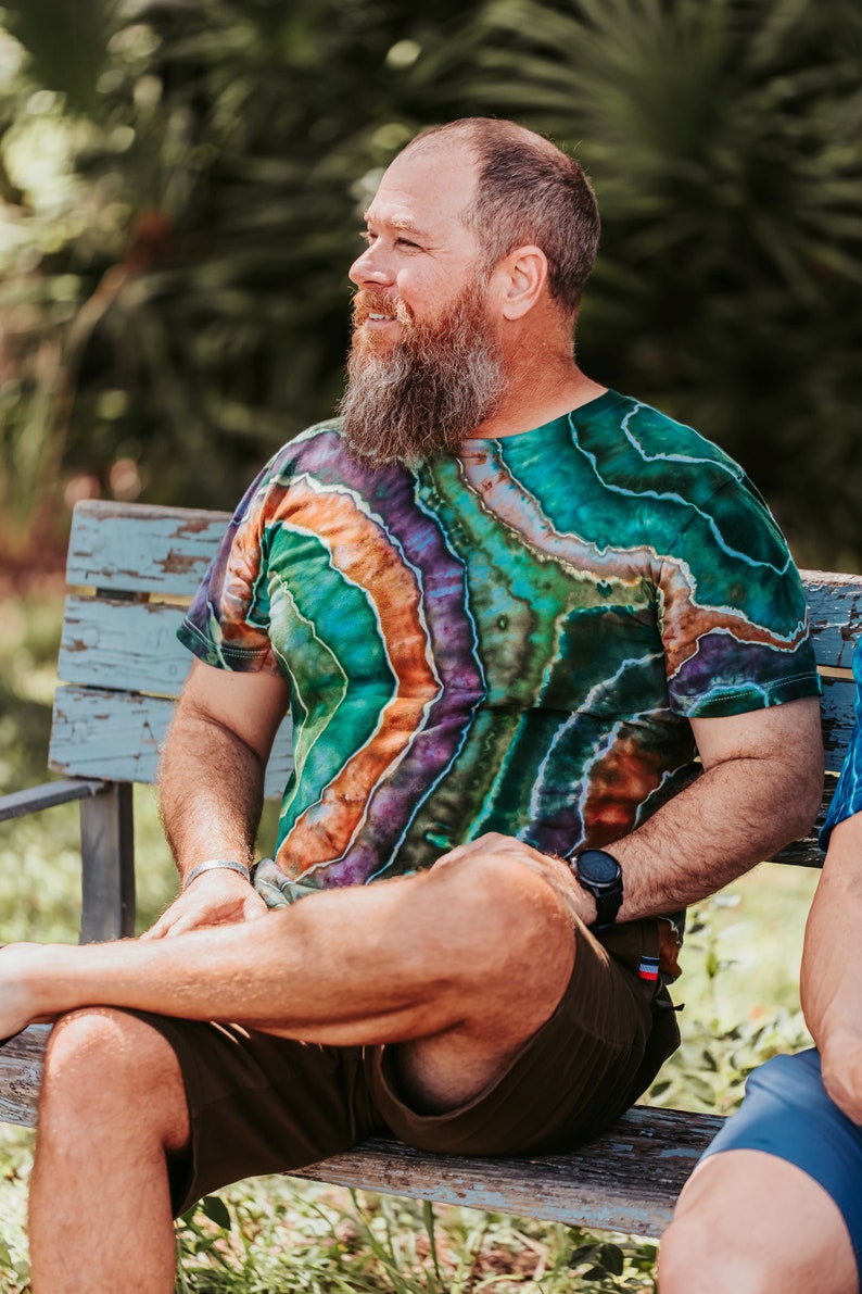 K&ouml;nnte beinhalten: Ein Mann mit langem, grauem Bart sitzt auf einer Holzbank und tr&auml;gt ein gr&uuml;n-orange-violettes Batik-Shirt und braune Shorts. Er tr&auml;gt eine Uhr am linken Handgelenk.
