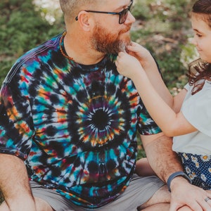 May include: A man with a beard and sunglasses sits cross-legged on the grass while a young girl gently strokes his beard. He is wearing a colorful tie-dye shirt and gray shorts. The girl is wearing a white shirt and a floral skirt.