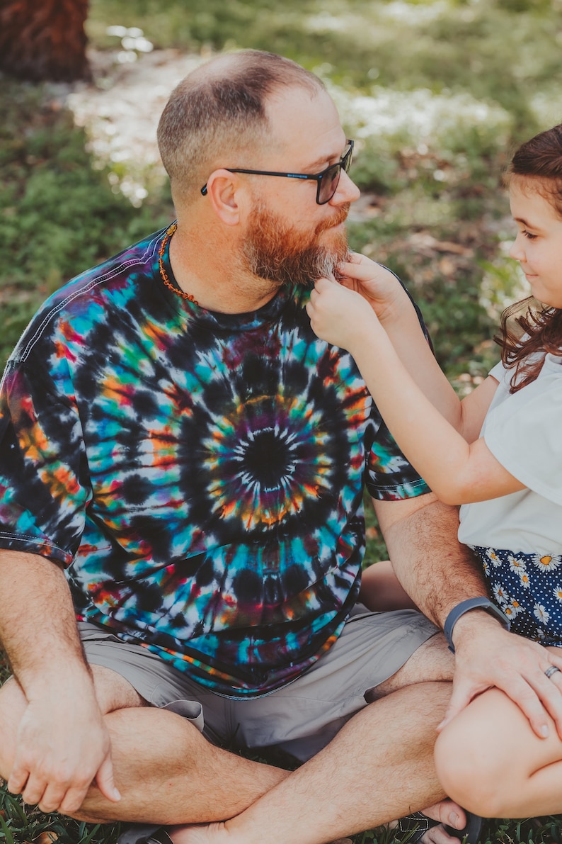 May include: A man with a beard and sunglasses sits cross-legged on the grass while a young girl gently strokes his beard. He is wearing a colorful tie-dye shirt and gray shorts. The girl is wearing a white shirt and a floral skirt.