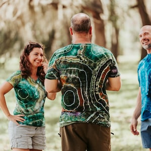 K&ouml;nnte beinhalten: Drei Personen in Batik-Shirts stehen in einem grasbewachsenen Bereich. Die Person in der Mitte tr&auml;gt ein gr&uuml;n-braun-violettes Batik-Shirt. Die Person links tr&auml;gt ein gr&uuml;n-gelbes Batik-Shirt. Die Person rechts tr&auml;gt ein blaues Batik-Shirt.