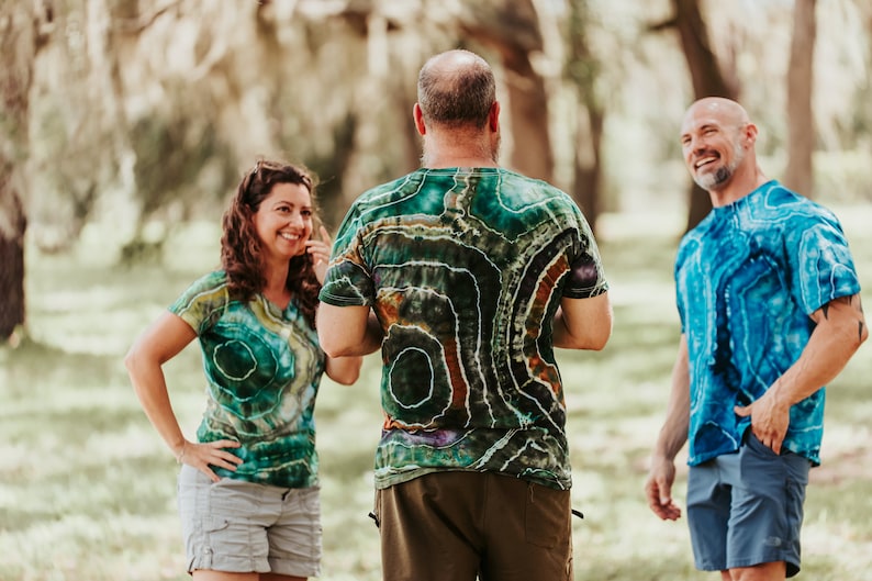 K&ouml;nnte beinhalten: Drei Personen in Batik-Shirts stehen in einem grasbewachsenen Bereich. Die Person in der Mitte tr&auml;gt ein gr&uuml;n-braun-violettes Batik-Shirt. Die Person links tr&auml;gt ein gr&uuml;n-gelbes Batik-Shirt. Die Person rechts tr&auml;gt ein blaues Batik-Shirt.