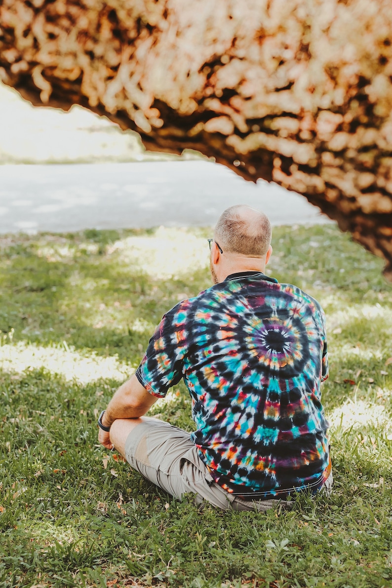 May include: A person wearing a colorful tie-dye shirt and khaki shorts sits on the grass with their back to the camera.