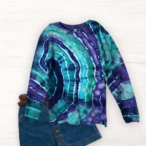 May include: A long-sleeve shirt featuring a tie-dye pattern in shades of deep purple, teal, and bright blue, arranged in a circular spiral design. The shirt is paired with a blue denim skirt that has a brown braided belt at the waistline.