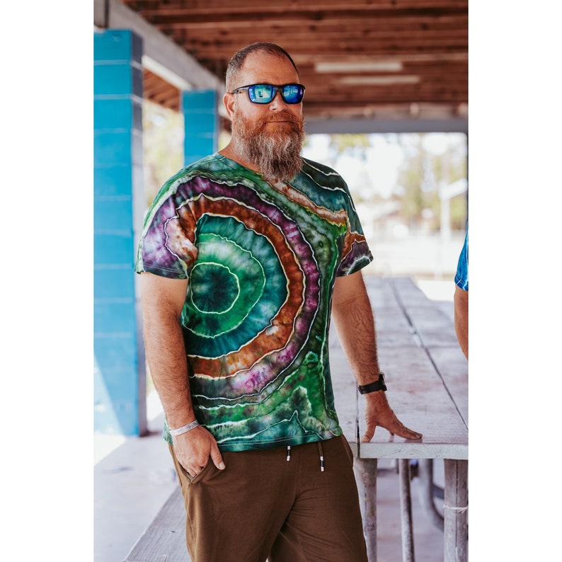 Bearded man with reflective sunglasses standing under a covered porch. he is wearing a geode tie dye shirt. It is a short sleeve cotton, ice dyed in greens, brown and purple.