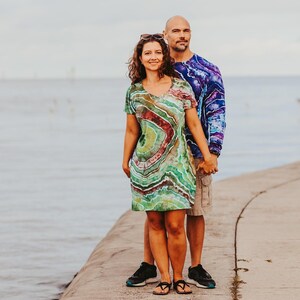 May include: A couple stands on a concrete pier. The woman wears a tie-dye dress with green, red, and brown swirls. The man wears a long-sleeved tie-dye shirt in blue and purple. They are holding hands.