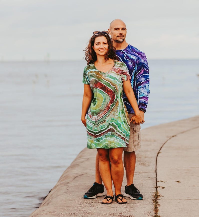 May include: A couple stands on a concrete pier. The woman wears a tie-dye dress with green, red, and brown swirls. The man wears a long-sleeved tie-dye shirt in blue and purple. They are holding hands.