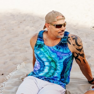 May include: A couple sitting on a beach blanket. The man is wearing a blue and green tie-dye tank top, a baseball cap, and sunglasses. The woman is wearing a pink and green tie-dye tank top and a straw hat.