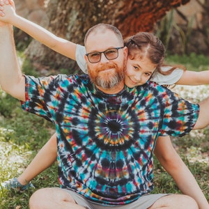 May include: A man wearing a colorful tie-dye shirt and grey shorts sits with a young girl on his lap. The girl is holding onto the man's arms and smiling at the camera. They are sitting in a grassy area with a large tree in the background.