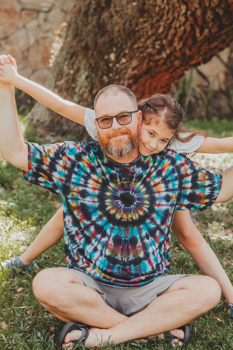 May include: A man wearing a colorful tie-dye shirt and grey shorts sits with a young girl on his lap. The girl is holding onto the man's arms and smiling at the camera. They are sitting in a grassy area with a large tree in the background.