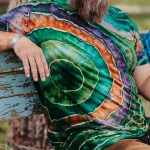 K&ouml;nnte beinhalten: Eine Person tr&auml;gt ein gr&uuml;n-orange-violettes Batik-Shirt mit einem kreisf&ouml;rmigen Muster. Das Shirt ist aus weichem, bequemem Material und hat eine lockere Passform.