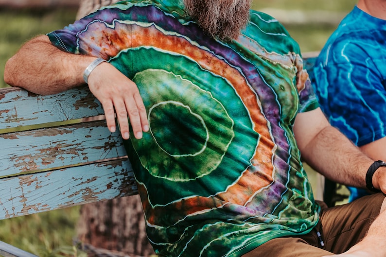K&ouml;nnte beinhalten: Eine Person tr&auml;gt ein gr&uuml;n-orange-violettes Batik-Shirt mit einem kreisf&ouml;rmigen Muster. Das Shirt ist aus weichem, bequemem Material und hat eine lockere Passform.