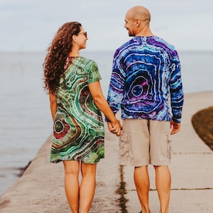 May include: A couple holding hands, seen from behind, on a concrete pier. The woman wears a green, brown, and red tie-dye dress. The man wears a blue and purple tie-dye long-sleeve shirt, khaki shorts, and black sneakers. The background is a body of water and a cloudy sky.