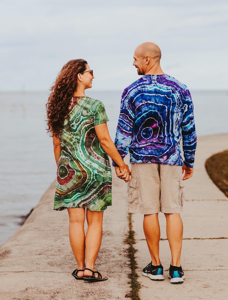May include: A couple holding hands, seen from behind, on a concrete pier. The woman wears a green, brown, and red tie-dye dress. The man wears a blue and purple tie-dye long-sleeve shirt, khaki shorts, and black sneakers. The background is a body of water and a cloudy sky.