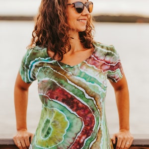 May include: A V-neck dress with a unique tie-dye pattern in shades of green, red, and blue. The dress has short sleeves and a fitted silhouette. The person is wearing sunglasses and is leaning on a wooden railing.