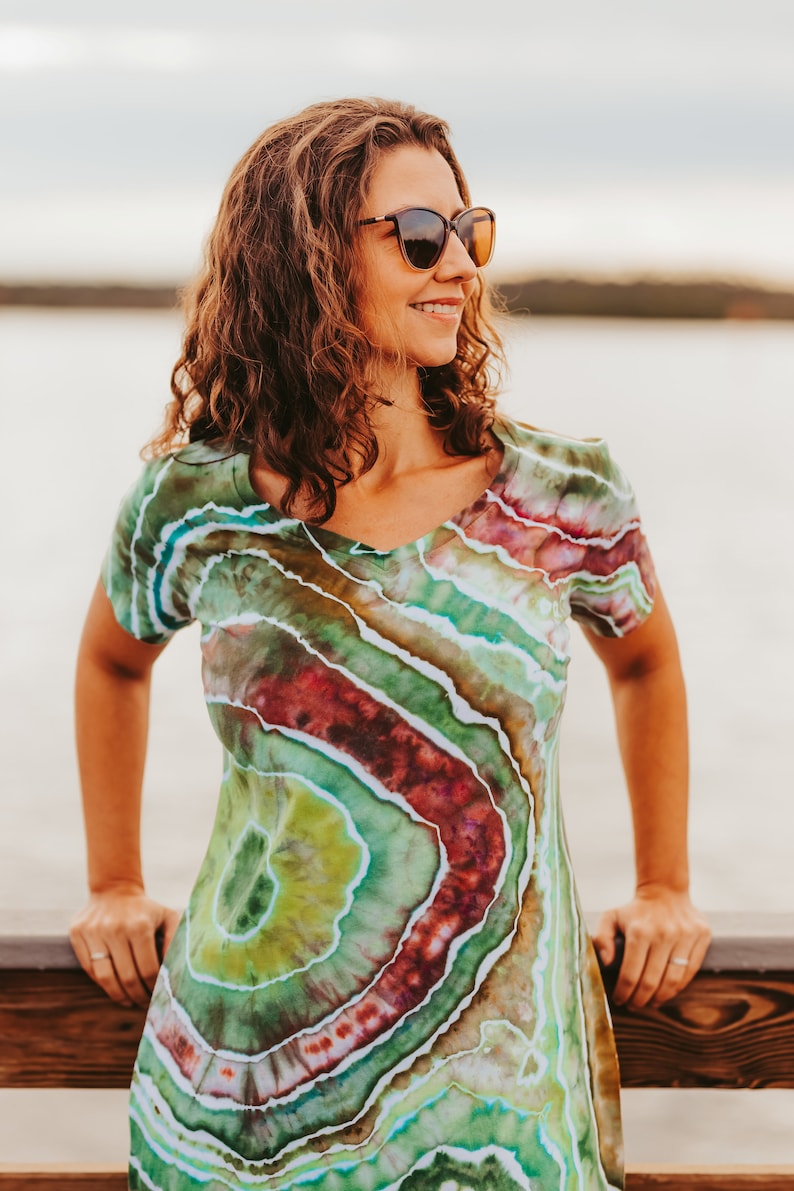 May include: A V-neck dress with a unique tie-dye pattern in shades of green, red, and blue. The dress has short sleeves and a fitted silhouette. The person is wearing sunglasses and is leaning on a wooden railing.