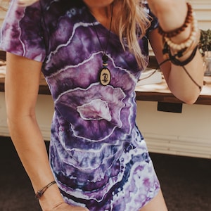 May include: A purple and white tie-dye dress with a V-neck. The dress features a swirling pattern and is accessorized with a wooden pendant necklace and multiple bracelets. The dress is knee-length.