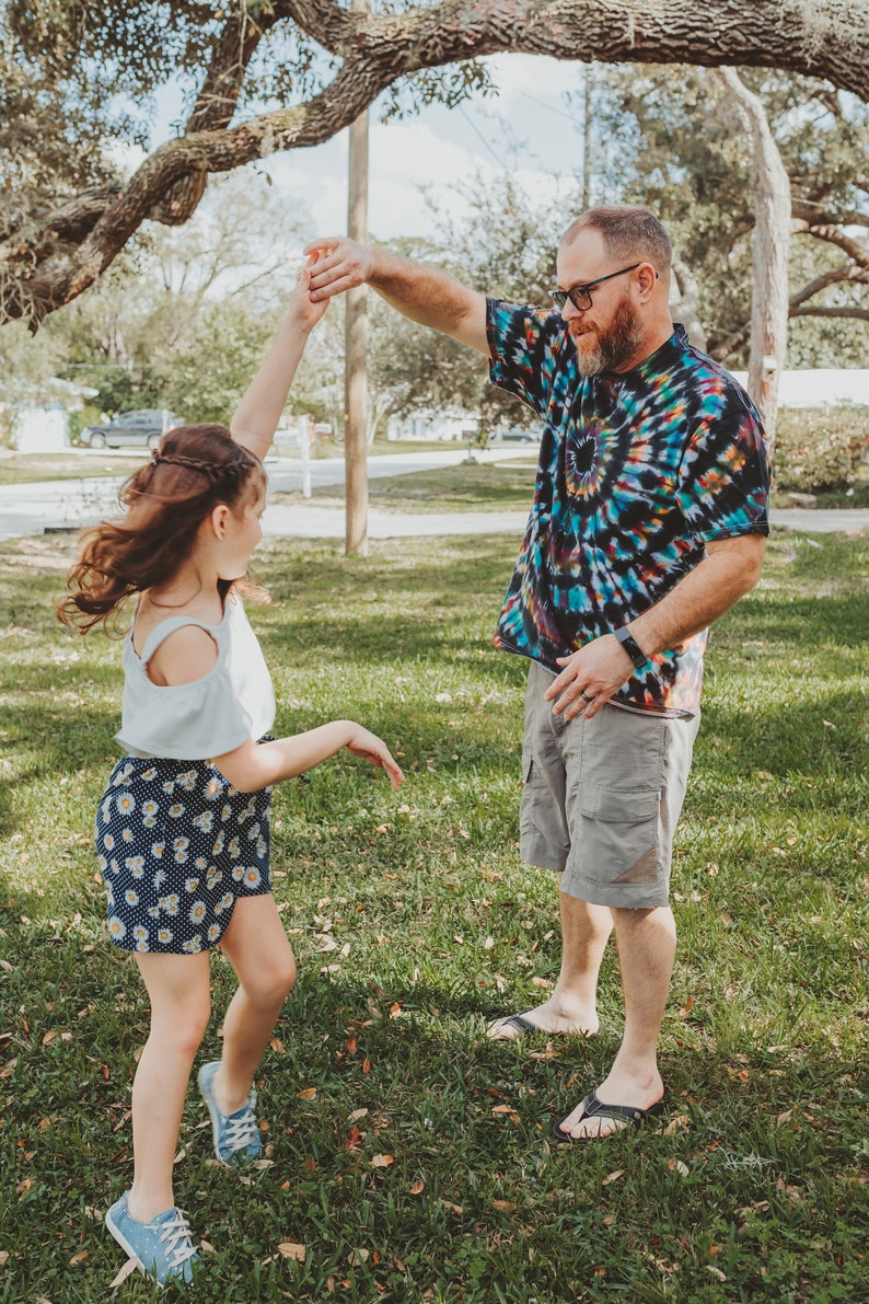 May include: A father and daughter are dancing in a grassy park. The father is wearing a tie-dye shirt and khaki shorts. The daughter is wearing a white top and a floral skirt. They are both smiling and having fun.