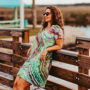 May include: A short-sleeved dress with a vibrant tie-dye pattern in shades of green, red, and brown. The dress has a V-neck and is knee-length. The person is wearing sunglasses and leaning against a wooden railing.