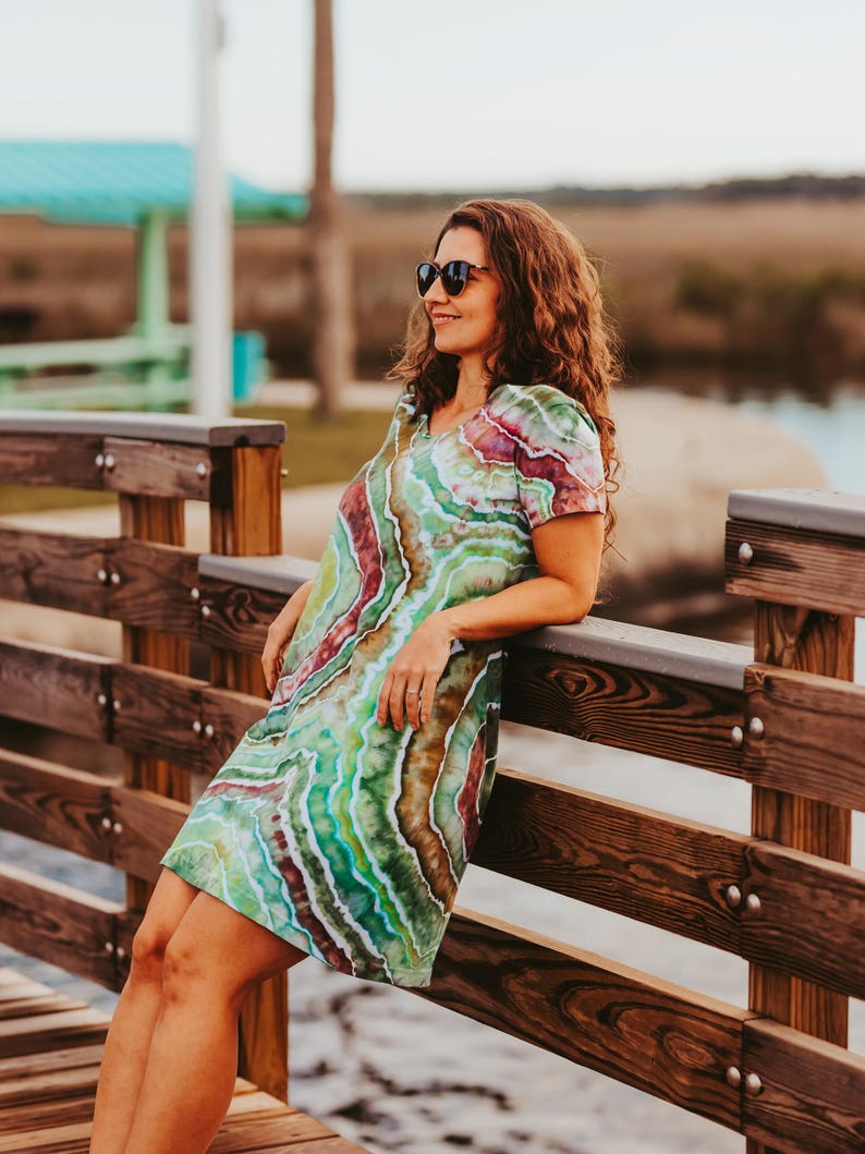May include: A short-sleeved dress with a vibrant tie-dye pattern in shades of green, red, and brown. The dress has a V-neck and is knee-length. The person is wearing sunglasses and leaning against a wooden railing.