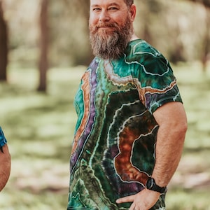 K&ouml;nnte beinhalten: Ein Mann mit Bart tr&auml;gt ein gr&uuml;n-lila-oranges Batik-Shirt mit einem braun-gr&uuml;n-schwarzen Muster. Er tr&auml;gt eine braune Hose und eine Uhr am linken Handgelenk.