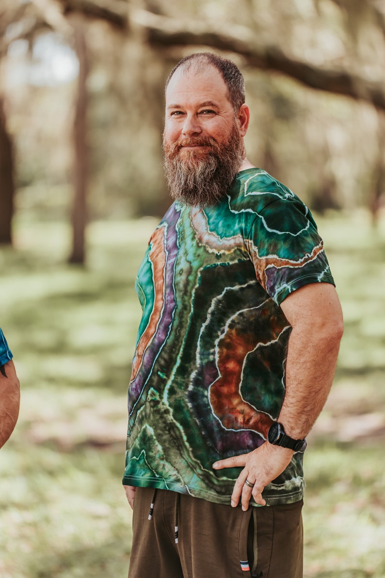 K&ouml;nnte beinhalten: Ein Mann mit Bart tr&auml;gt ein gr&uuml;n-lila-oranges Batik-Shirt mit einem braun-gr&uuml;n-schwarzen Muster. Er tr&auml;gt eine braune Hose und eine Uhr am linken Handgelenk.