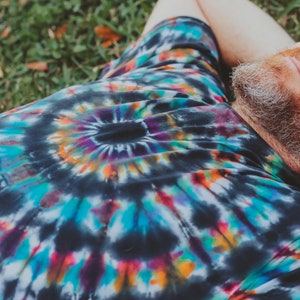 May include: A person wearing a black tie-dye shirt with a colorful swirl pattern. The person is lying on their back on a bed of green grass.
