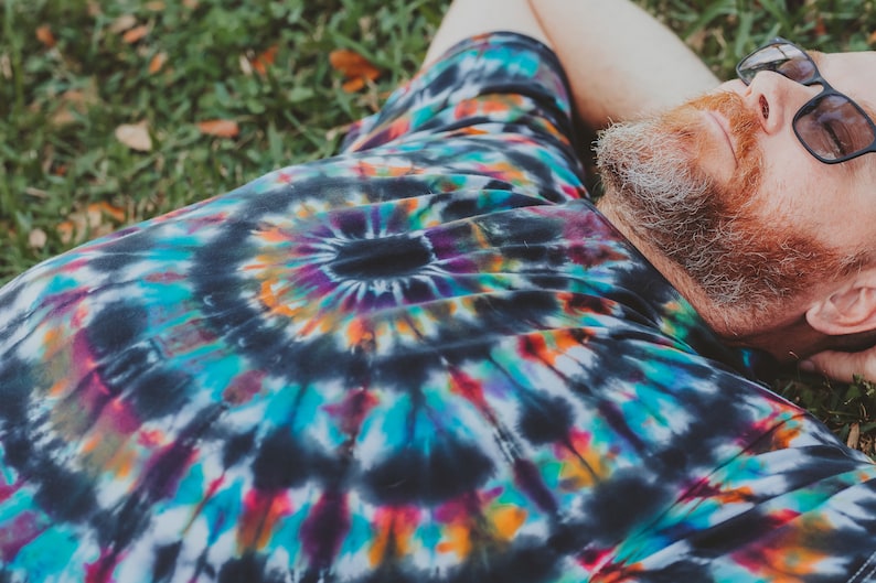 May include: A person wearing a black tie-dye shirt with a colorful swirl pattern. The person is lying on their back on a bed of green grass.