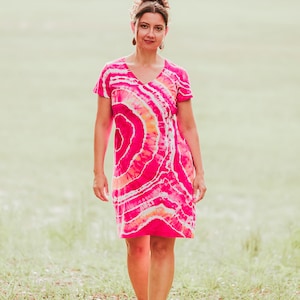 May include: A vibrant, short-sleeved dress with a V-neck, featuring a tie-dye pattern in shades of pink, orange, and white. The dress is knee-length and worn outdoors. The overall theme is casual summer wear.