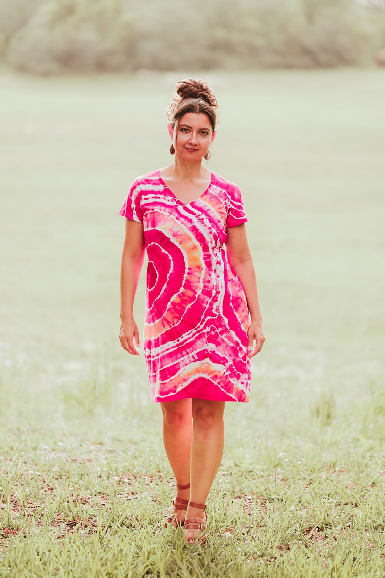 May include: A vibrant, short-sleeved dress with a V-neck, featuring a tie-dye pattern in shades of pink, orange, and white. The dress is knee-length and worn outdoors. The overall theme is casual summer wear.