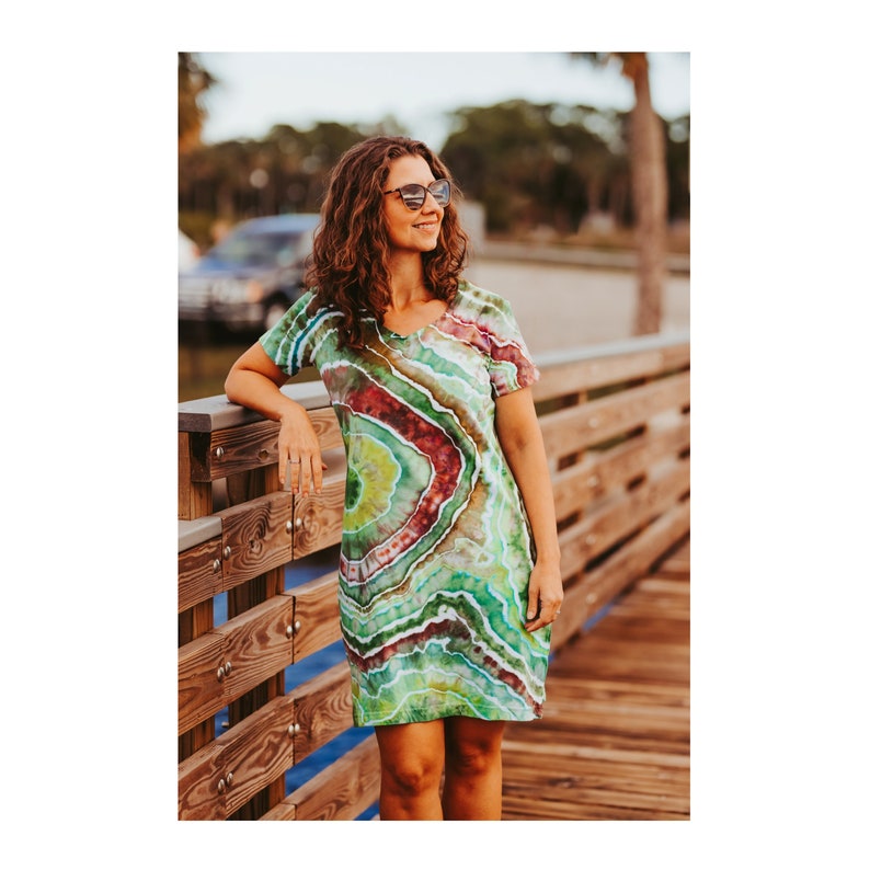 May include: A short-sleeved, V-neck dress with a vibrant tie-dye pattern in shades of green, red, and white. The dress has a fitted silhouette and falls to mid-thigh. The tie-dye design creates a layered, agate-like effect. The dress is worn outdoors.