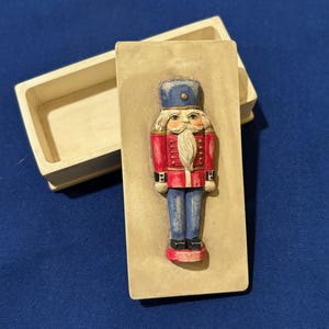 May include: A wooden Christmas ornament in the shape of a nutcracker soldier. The soldier wears a blue hat, red jacket, and blue pants. The ornament is inside a rectangular box with a cream-colored finish.