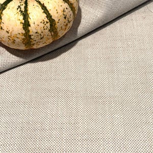 May include: A small, decorative gourd with a cream-colored base and green stripes rests on a textured, light gray fabric. The gourd's stem is visible, and the fabric is folded, creating shadows.