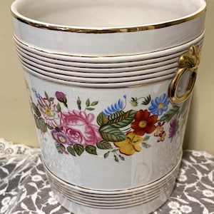 May include: A white ceramic ice bucket with a crackle glaze finish, featuring a colorful floral pattern of pink, red, yellow, and blue flowers. The bucket has gold-toned horizontal ribbed bands at the top and bottom and gold-toned ring handles.