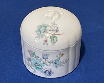 May include: A white ceramic trinket box with a lid, adorned with a floral pattern in shades of blue, green, and pink. The box has a rounded shape with a ribbed design on the sides and a small floral accent on the lid.