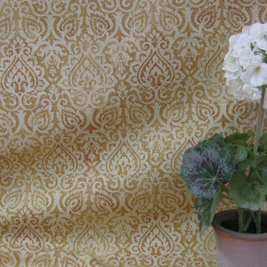 May include: A white flower with green leaves in a brown pot against a background of a beige fabric with a brown damask pattern.