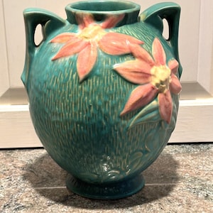 May include: A teal ceramic vase with two handles and embossed floral designs. The vase features two large, pink and yellow flowers. The vase has a textured surface and a round base, suitable for home decor.