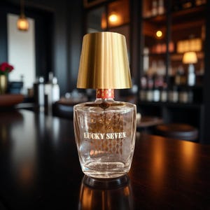 May include: A unique table lamp made from a clear glass bottle with the text "LUCKY SEVEN" and a gold lampshade. The lamp has a red and gold accent at the base of the lampshade. The lamp is on a dark surface.