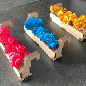 May include: Three wooden flower boxes with different colored paper flowers. The boxes are painted white and have a brown stain on the edges. The flowers are blue, pink, and yellow.