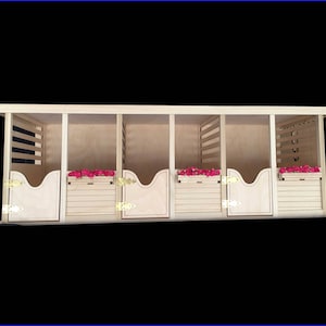 May include: A wooden miniature horse stable with four stalls, each with a hinged door and a hay rack. The stalls are painted a light brown color and have a white trim. The stable is set against a black background.
