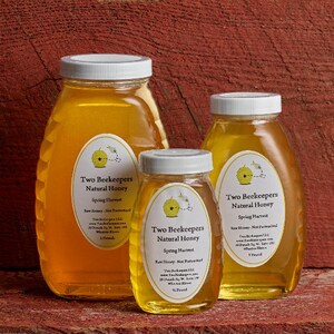 May include: Three glass jars of honey with labels that read "Two Beekeepers Natural Honey" and "Spring Harvest". The jars are on a red wooden surface.