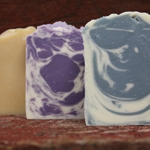 May include: Three rectangular bars of handmade soap. The first is a pale yellow, the second has a purple and white swirl pattern, and the third has a blue and white swirl pattern. The soaps are displayed on a rustic, reddish-brown wooden surface.