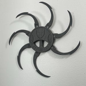 Könnte beinhalten: Graues, sternförmiges Dekorationsobjekt mit gebogenen, klauenartigen Armen, die sich von einer zentralen, kreisförmigen Nabe aus erstrecken. Die Nabe weist ein detailliertes, strukturiertes Design auf. Das Objekt ist an einer weißen Wand befestigt.