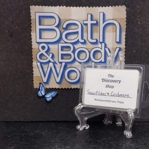 May include: A clear plastic container with a white label that reads "The Discovery Shop Snowflakes & Cashmere Hand poured for you. Enjoy" on a black surface.
