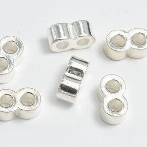 Peut inclure: Plusieurs perles en métal argenté. Chaque perle a la forme d'un huit, avec deux trous circulaires. Les perles ont une surface lisse et brillante, reflétant la lumière. Elles sont probablement destinées à la fabrication de bijoux ou à l'artisanat.