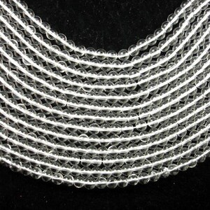 May include: A strand of clear round glass beads, arranged in a curved pattern. The beads are smooth and translucent, with a slight sparkle.