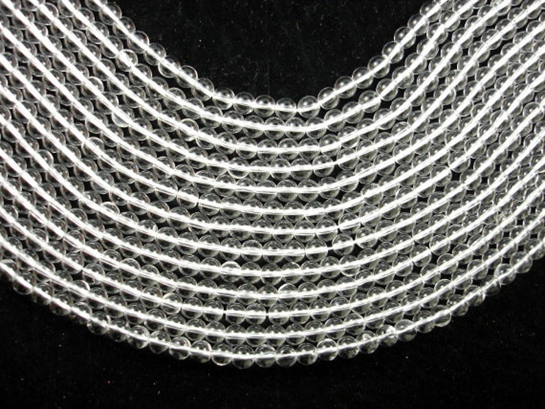 May include: A strand of clear round glass beads, arranged in a curved pattern. The beads are smooth and translucent, with a slight sparkle.