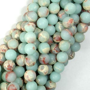 May include: A strand of round, light blue and white marbled gemstone beads. The beads have a mottled, speckled appearance with hints of red.