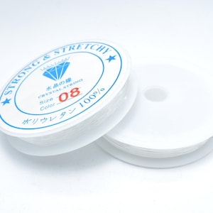 Peut inclure: Deux bobines de fil de cristal transparent et extensible. L'une des bobines est partiellement déroulée. L'étiquette indique "STRONG & STRETCHY", "CRYSTAL STRING", "Size 08" et "100%" en anglais et en japonais.