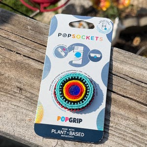 May include: A circular phone grip featuring a concentric beaded pattern in bright turquoise, black, yellow, and red, with a deep blue center. The accessory is mounted on a white and blue branded PopSockets packaging card with plant-based material text.